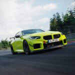 2027 BMW M2 coupe in Velvet Blue driving on mountain road, showcasing aggressive M front grille, quad exhaust, and carbon fiber details