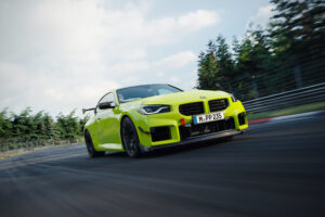 2027 BMW M2 coupe in Velvet Blue driving on mountain road, showcasing aggressive M front grille, quad exhaust, and carbon fiber details